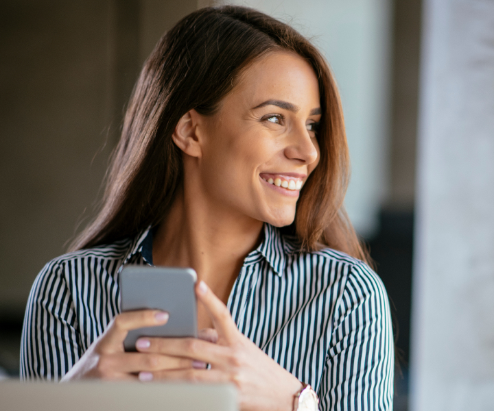 Mulher olhando para o lado e sorrindo segurando um smarphone na altura do rosto.