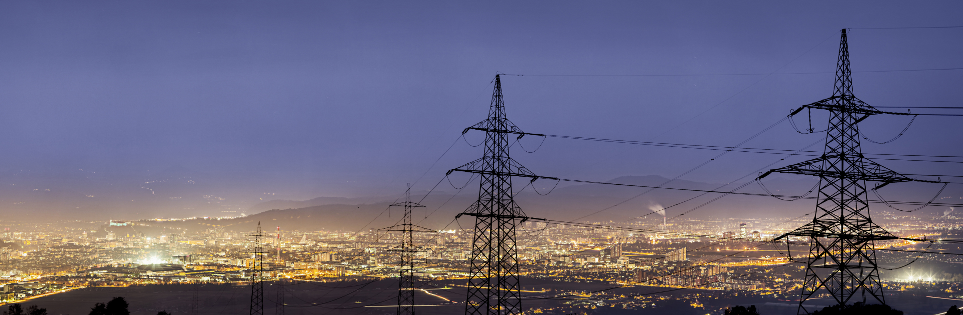 Imagem panoramica de uma cidade iluminada, com postes de alta voltagem em primeiro plano.